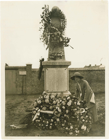 Black and white photograph of a woman in 1920s clothing placing a large floral wreath at the foot of a small stone obelisk with the inscription 'Gloire et Honneur'.