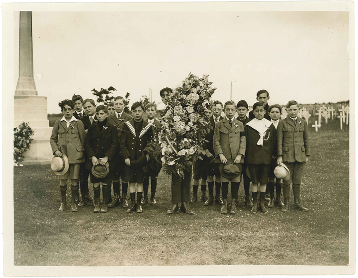 Black and white photograph of a group of boys in school uniform, one in centre holding a large floral wreath. To the left a small stone obelisk and to the right rows of white cross grave markers.
