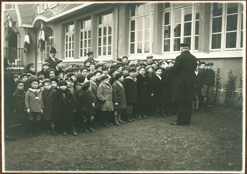 Black and white photograph of a large group of children in coats and caps standing outside a school singing. A man in a cap stands facing them. Two men stand behind them, one playing a clarinet.