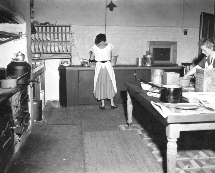 Black and white photograph of two women working in a commercial kitchen, to one side a wood stove.