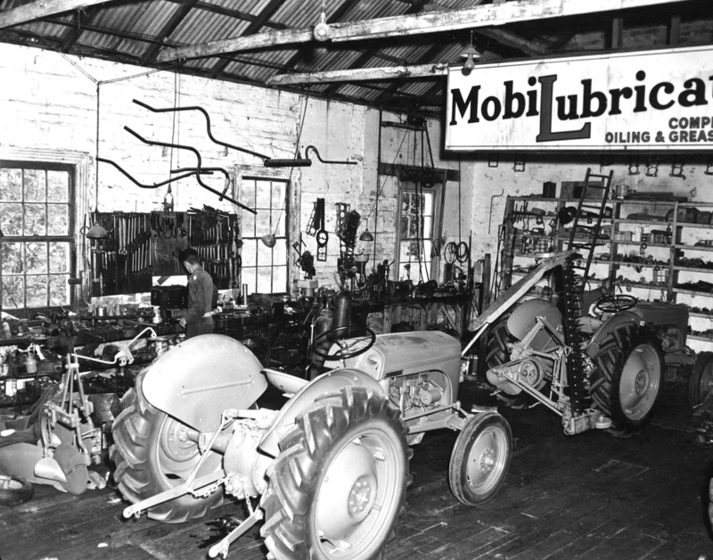 Black and white photograph of the interior of a motor mechanics workshop with a tractor, shelves and tools on the wall and a person at a bench.