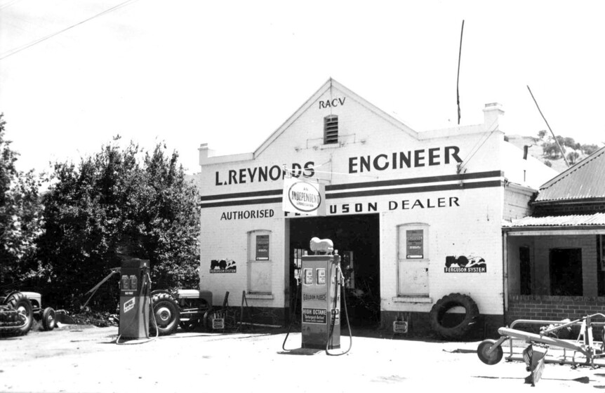 Tractor and two petrol bowsers outside a white shop front with gable and central garage door. Signage says, 'L. Reynolds Engineer.'