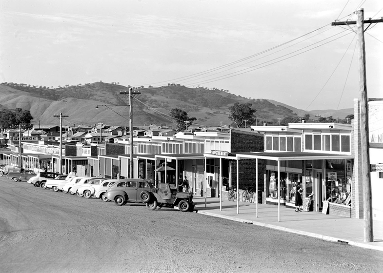 Black and white photograph of a row of 1950s cars angle parked outside a row of 1950s shopfronts. Hills in distance.