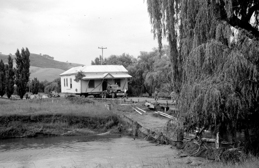 Black and white photograph of a single storey weatherboard house on a truck approaching a small bridge. Stream in foreground. Hills in background.