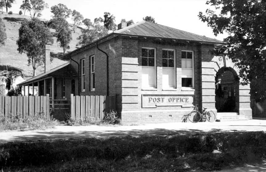 Black and white photograph of a single storey red brick building with an iron hip roof, triple windows at the front and an arched entrance. Sign says, 'Post Office.'