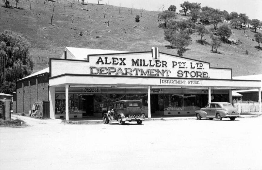 Black and white photograph of a single storey shopfront with a deep veranda and two cars parked outside. Sign says, 'Alex Miller Pty. Ltd. Department Store.'