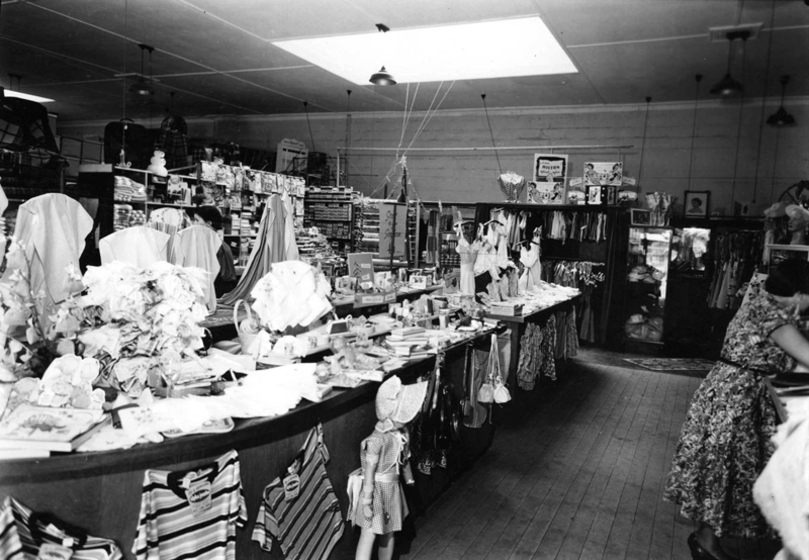 Black and white photograph of the interior of a shop with table linen, clothing and a doll.