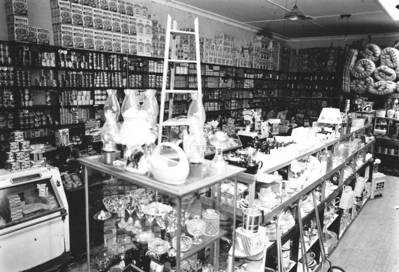 Black and white photograph of the interior of a shop with crockery, packaged food and mattresses.