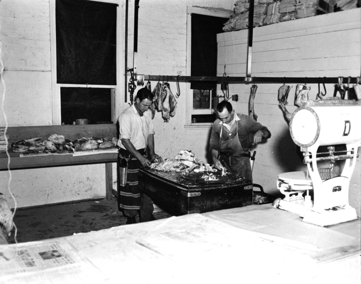 Black and white photograph of the interior of a butcher shop with two men in aprons cutting up meat. Carcases hanging from hooks and scales in the foreground.