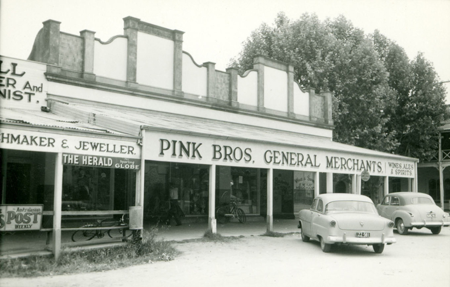 Black and white photograph of a single storey shopfront with a deep veranda with two cars parked outside. Sign says, 'Pink Bros. General Merchants.'