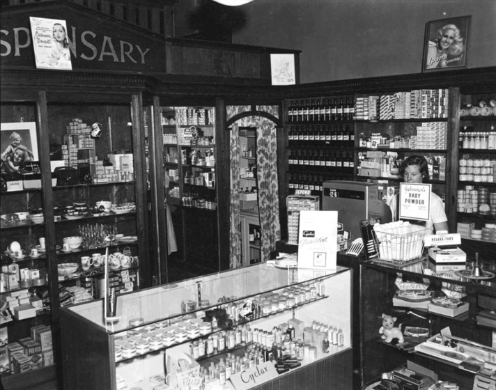 Black and white photograph of the interior of a pharmacy with well-stocked shelves and counters and a woman standing behind a cash register. Sign says, 'Dispensary.'