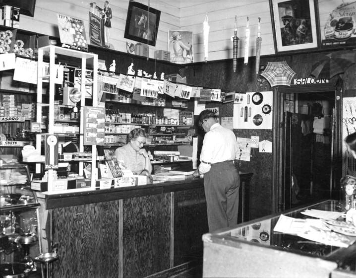 Black and white photograph of the interior of a well-stocked shop with a woman behind the counter and a man in front of the counter and the sign, 'Saloon', above the door.