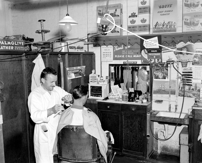 Black and white photograph a barber shop with a barber cutting a man's hair and advertisements covering the wall.