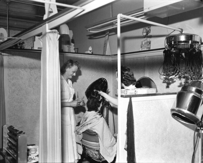 Black and white photograph of a hairdressing salon with two women cutting a woman's hair.