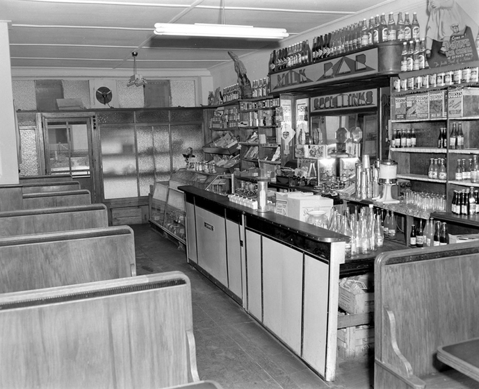 Black and white photograph of a small café with booths on the left and counter on the right. The shelves behind the counter are well-stocked with bottles of soft-drink.