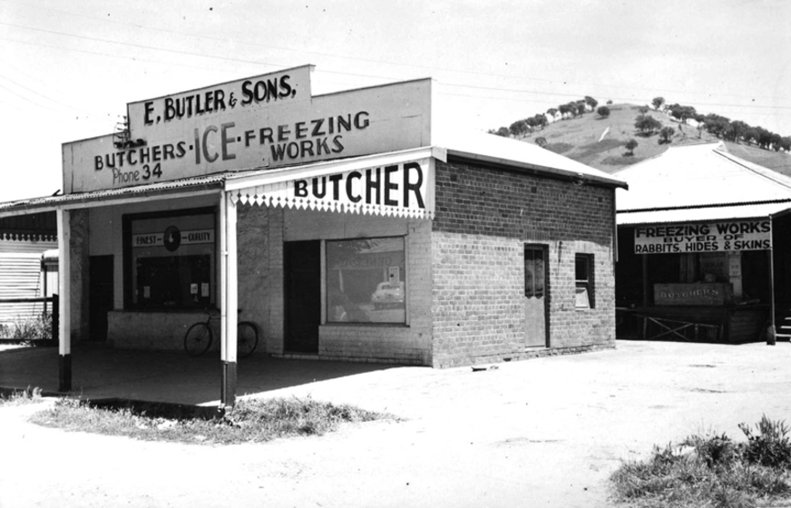 Black and white photograph of a small shop with a deep veranda. Sign says, 'E. Butler & Sons Butcher, Ice Freezing Works.'