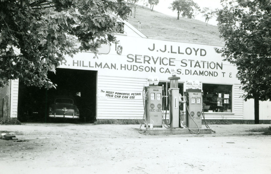 Black and white photograph of a single storey weatherboard building with an open garage door and two petrol bowsers in front. The sign says, 'J.J. Lloyd Service Station.'