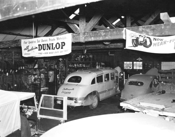 Black and white photograph of the interior of a mechanics workshop with five vehicles.