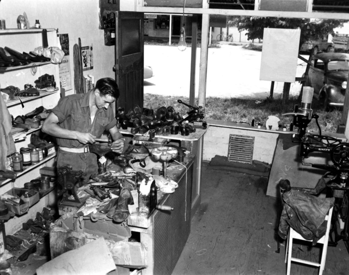 Black and white photograph of the interior of a cobbler's shop with a man behind a counter covered in shoes repairing a boot.