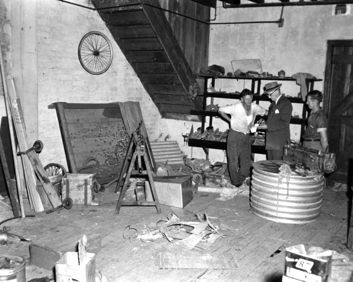 Black and white photograph of the interior of a workshop. Three men talking, one in a suit. Small corrugated water tank and metal offcuts on floor.