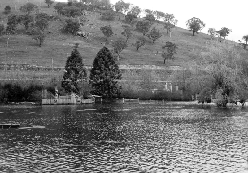 Black and white photograph of a body of water at the foot of a hill. From the water emerges a wooden building and trees.