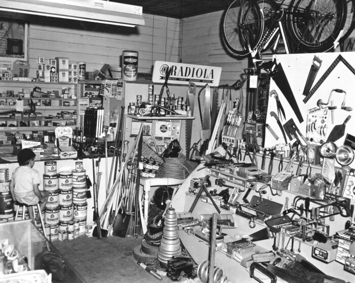 Black and white photograph of a small boy sitting on a stool at the counter of a well-stocked hardware store.