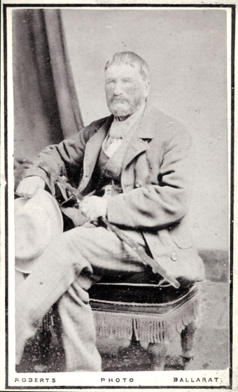Black and white studio photo portrait of a seated bearded middle-aged man wearing a Victorian suit and holding a hat and walking cane.