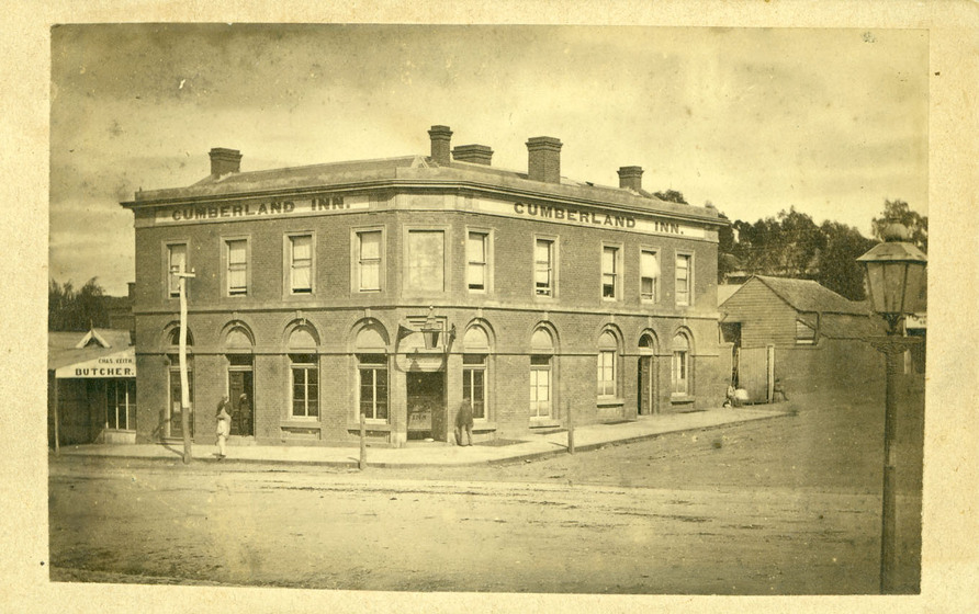 Black and white photograph of a two storey Victorian building on a corner with arched windows on the ground floor and chimneys. Signage says, 'Cumberland Inn'.