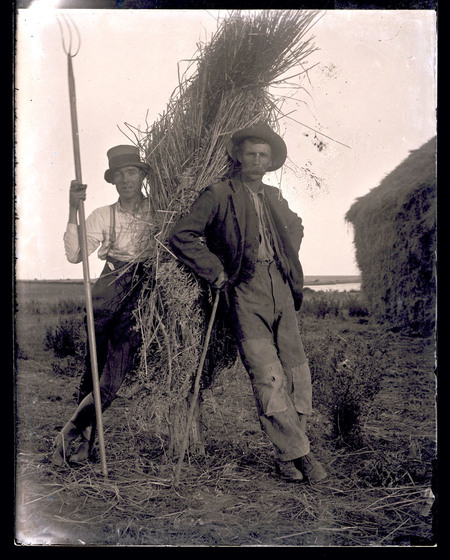 Black and white early Twentieth Century photograph of two labourers leaning on a sheaf, one holding a pitchfork, the other with a moustache.