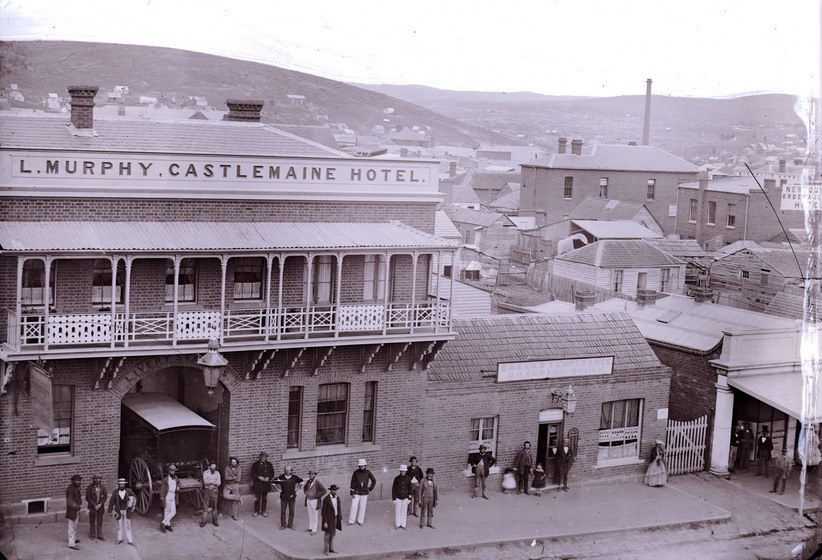Black and white photograph of people standing outside a Victorian era double storey brick building with a cart in the carriageway and a first floor veranda, the signage saying, 'L. Murphy, Castlemaine Hotel'. Taken from a high vantage point with a township in the distance.