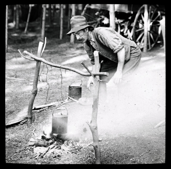 Black and white photograph of a tramp wearing a hat and moustache tending a can hanging over a small fire from a frame of branches in a bushland setting.