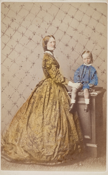 Black and white hand-tinted studio photo portrait of a woman in a large Victorian gown with her hand on the knee of a small boy sitting on a pillar.