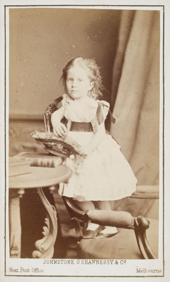 Sepia studio photo portrait of a small girl in a Victorian dress kneeling on a chair holding a basket beside a table with books on it.