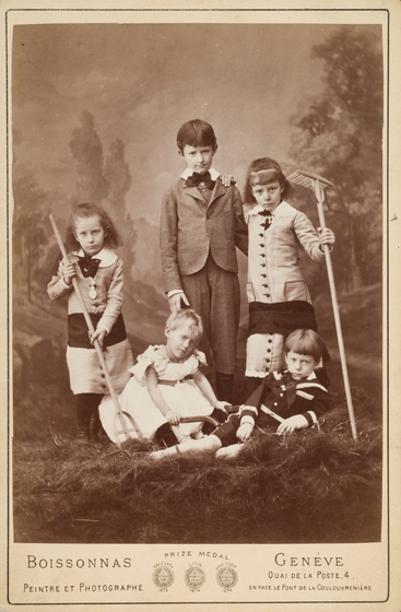 Sepia studio photo portrait of three girls and two boys well-dressed in Victorian clothing standing and sitting on straw, one holding a pitchfork and the another a rake.