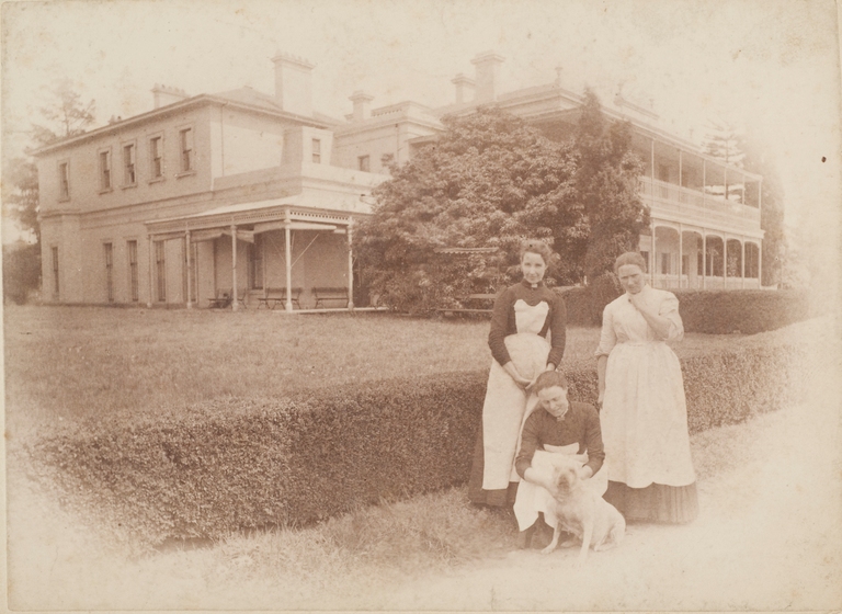 Sepia photograph of three maids in Victorian clothing, two standing and one squatting to pat a dog. Behind them a two storey Victorian mansion with verandahs on two floors.