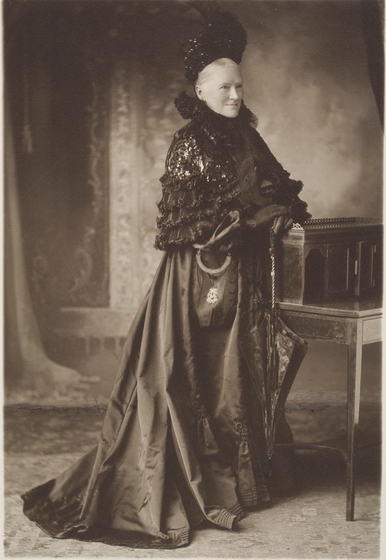 Black and white full length studio photo portrait of a woman in a long black Victorian dress, short sequined cape and headpiece, carrying an umbrella and leaning on a secretaire. Perhaps dressed in mourning.