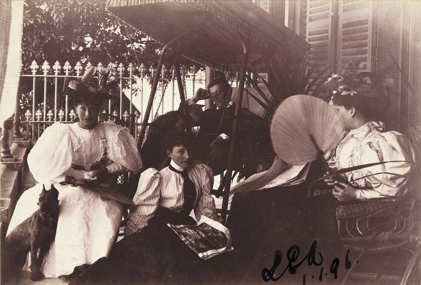 Black and white photograph of three young women and a young man in Victorian clothing sitting on a verandah with a dog. One woman carries a fan.
