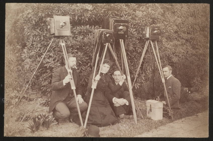 Sepia photograph of a woman and three men in Victorian clothing kneeling under four cameras on tripods in front of a bush.
