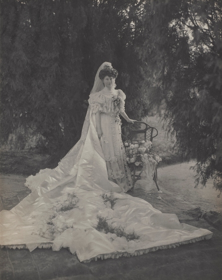 Black and white photograph of a woman in an elaborate Edwardian wedding gown with a lace bodice and an extensive train, standing under trees beside a cane chair with a bouquet on it.