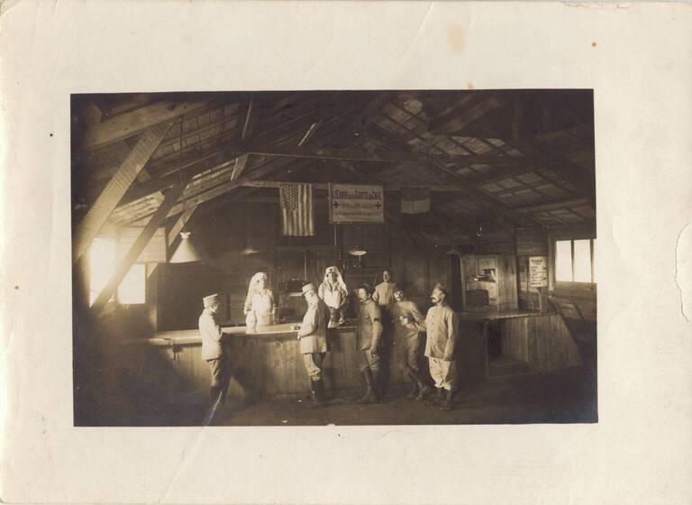 Black and white photograph of two women in Red Cross uniforms and a man standing behind a counter in a wooden hall. In front of the counter stand five men in French military uniforms. The sign above them says, 'l'Oeuvre de la Goutte de Café'.