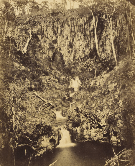 Sepia photograph of a wooded escarpment with with a waterfall flowing down to a pool.