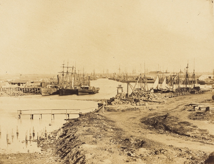Sepia photograph of an urban river with bare banks lined with warehouses and Nineteenth Century steam and sailing vessels.