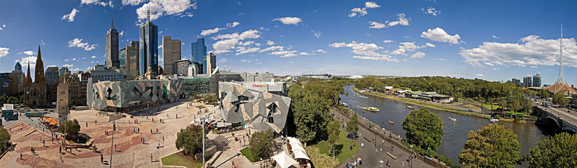 Colour photograph of a cityscape, a public square surrounded by lob-sided buildings, a river with boats and parkland under a blue sky with intermittent cloud cover.