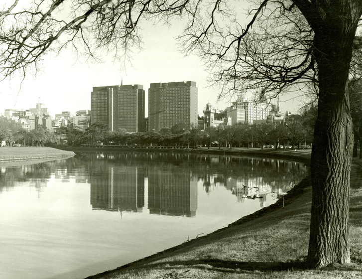 Black and white photograph of a city scape with a pair of modernist multi-storey buildings reflected in river.