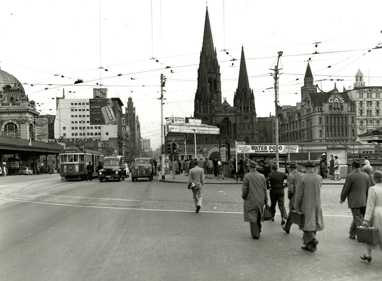 Black and white photograph of pedestrians crossing a city intersection, cathedral in distance.