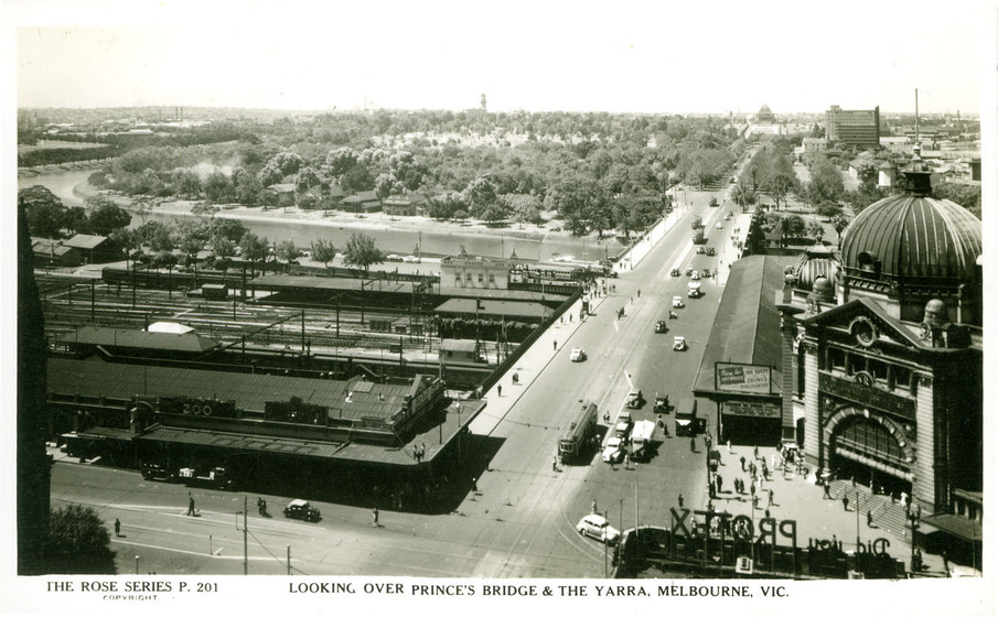 Black and white postcard of a city from a high vantage point showing a railway station, river, bridge and parkland beyond.