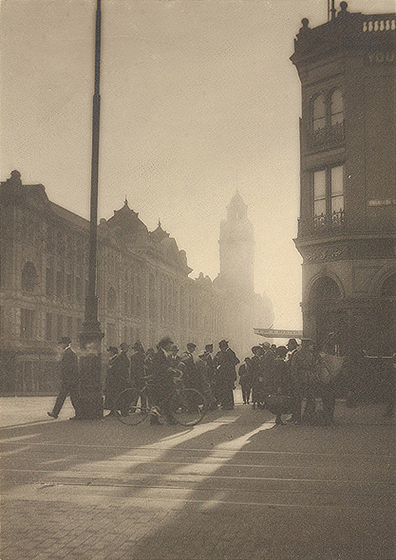 Black and white photograph of a city intersection with low sun shining through fog. Crowd on the corner in silhouette. Large Victorian building beyond.