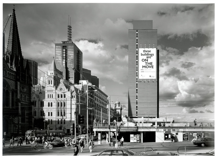 Black and white photograph of a city street. On the left is the spire of a church on the right is a modernist building with a banner on it reading, 'These Buildings are on the Move'.