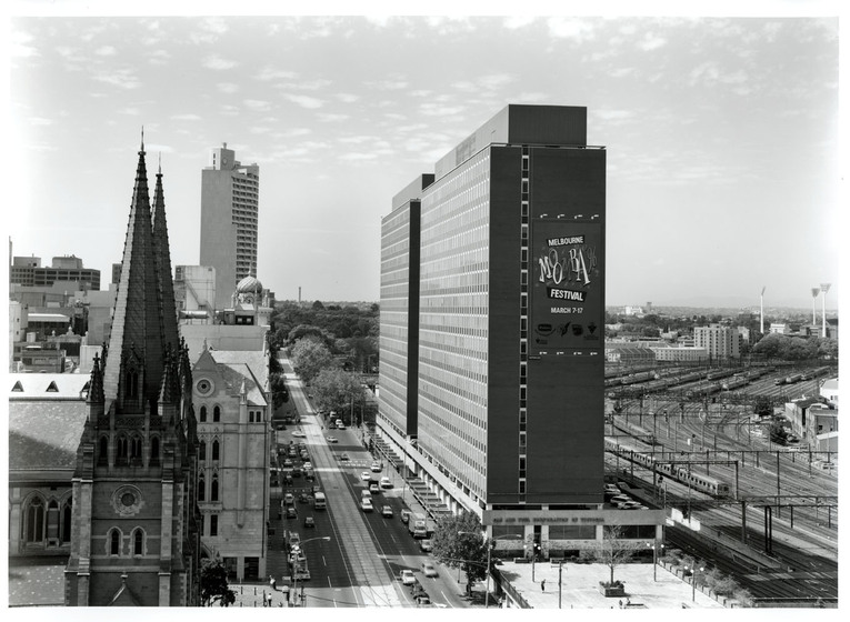 Black and white photograph of a city street from a high vantage point. On the left is the spire of a church on the right is a modernist building with a banner on it reading, 'Melbourne Moomba Festival March 7-17'. Railyards beyond.