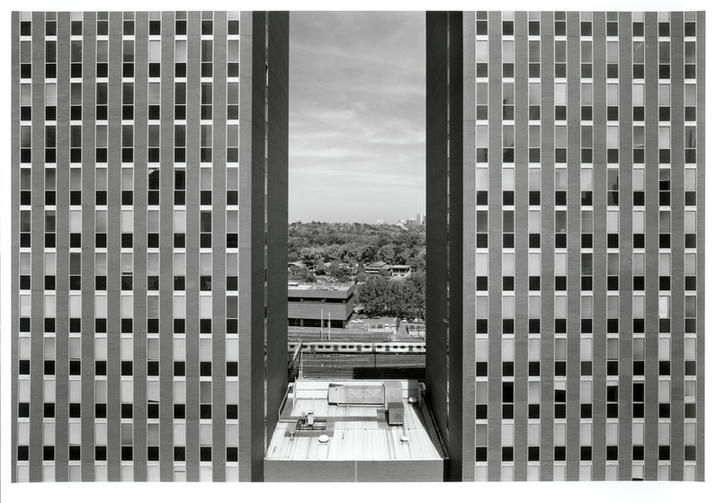 Black and white photograph of a close up looking between two modernist multi-storey buildings to parkland beyond.